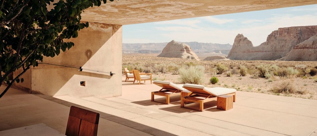 Two wooden loungers and a side table on the shaded terrace of an Amangiri suite, looking out over desert scrub to the sandstone mesas, with a pair of armchairs behind them and a tree at the left edge.
