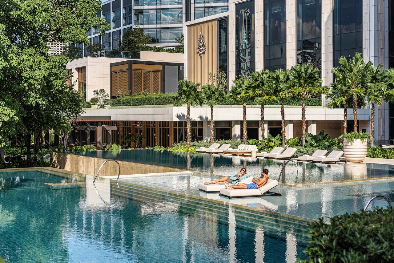 Two guests on a floating lounger in the infinity pool at Four Seasons Hotel Bangkok, palm trees and the hotel's tower behind them.