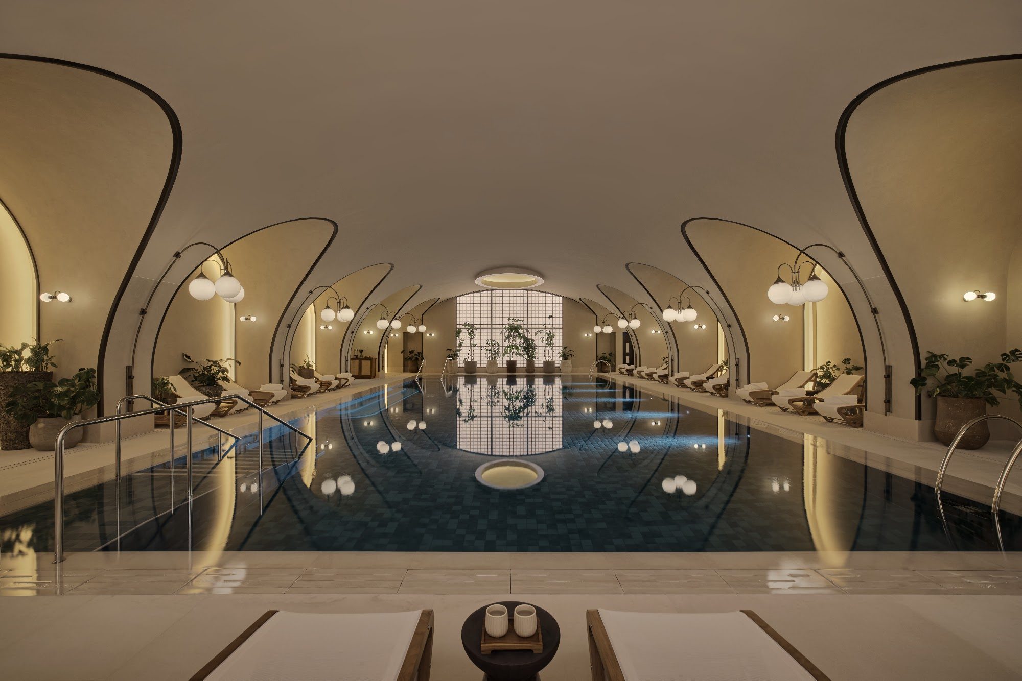 The indoor pool at Six Senses London, a long tiled pool under pale vaulted arches, with globe lamps and loungers reflected in still water.