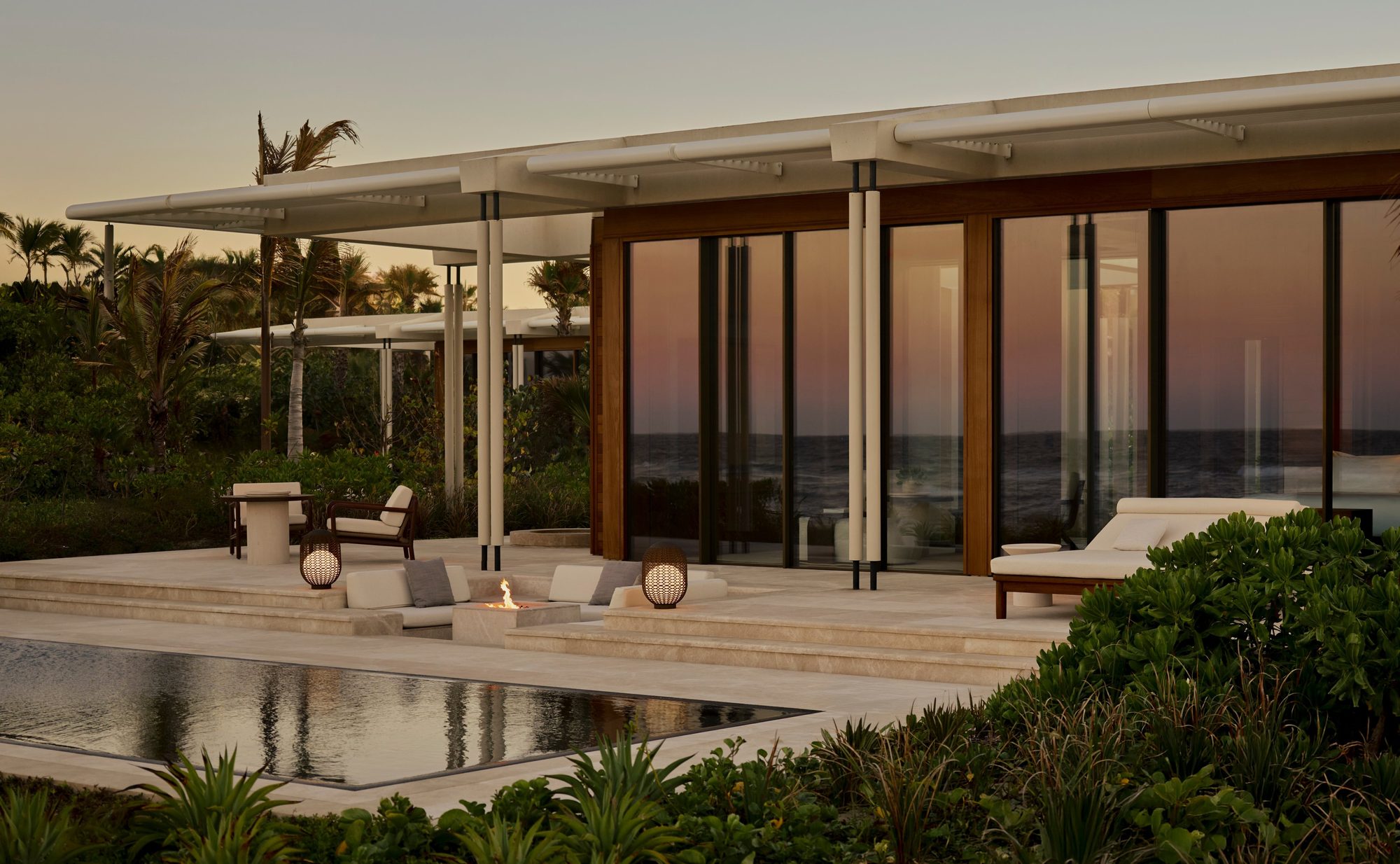 A poolside pavilion at Amanvari, Aman's Los Cabos resort, with an infinity pool, a fire pit lounge, and glass doors framing the sunset over the Pacific.