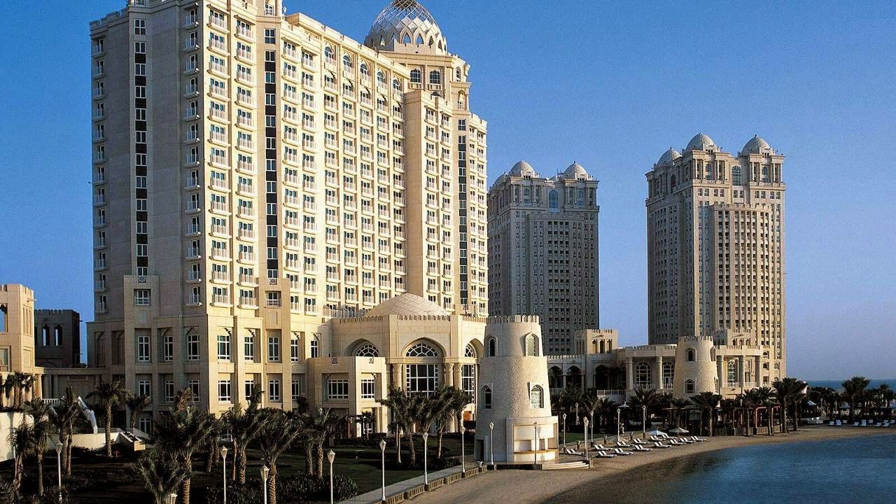 The cream towers of Four Seasons Hotel Doha rising over palm trees and a sandy beach lined with loungers.