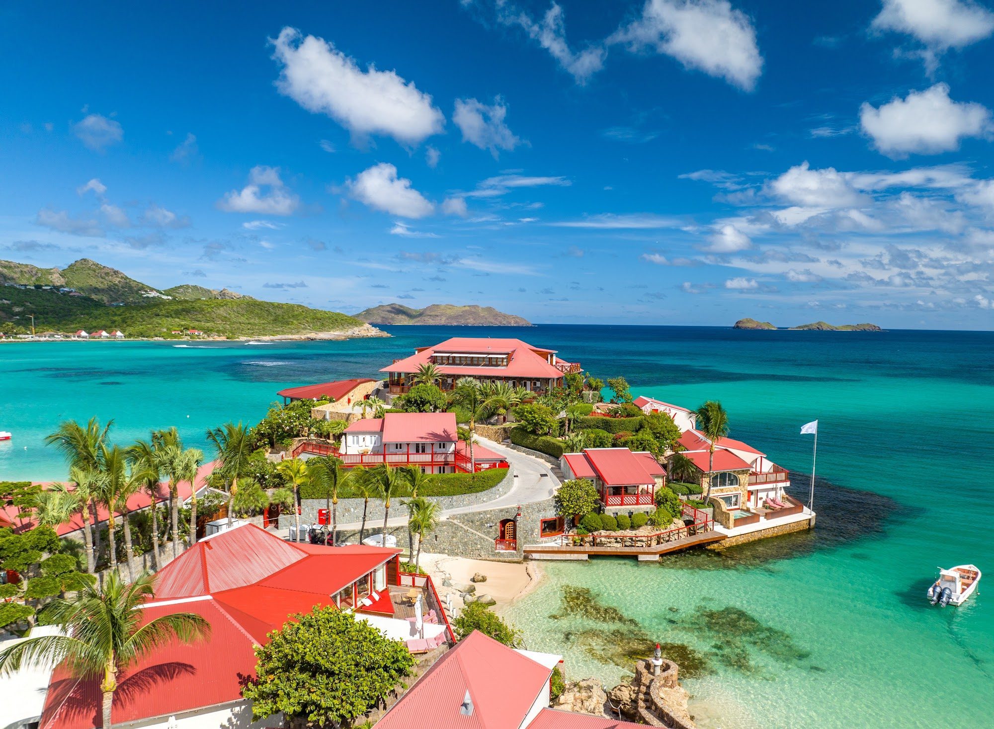 Aerial view of Eden Rock St Barths, red-roofed villas on a rocky peninsula surrounded by turquoise sea, with green hills and scattered clouds behind.