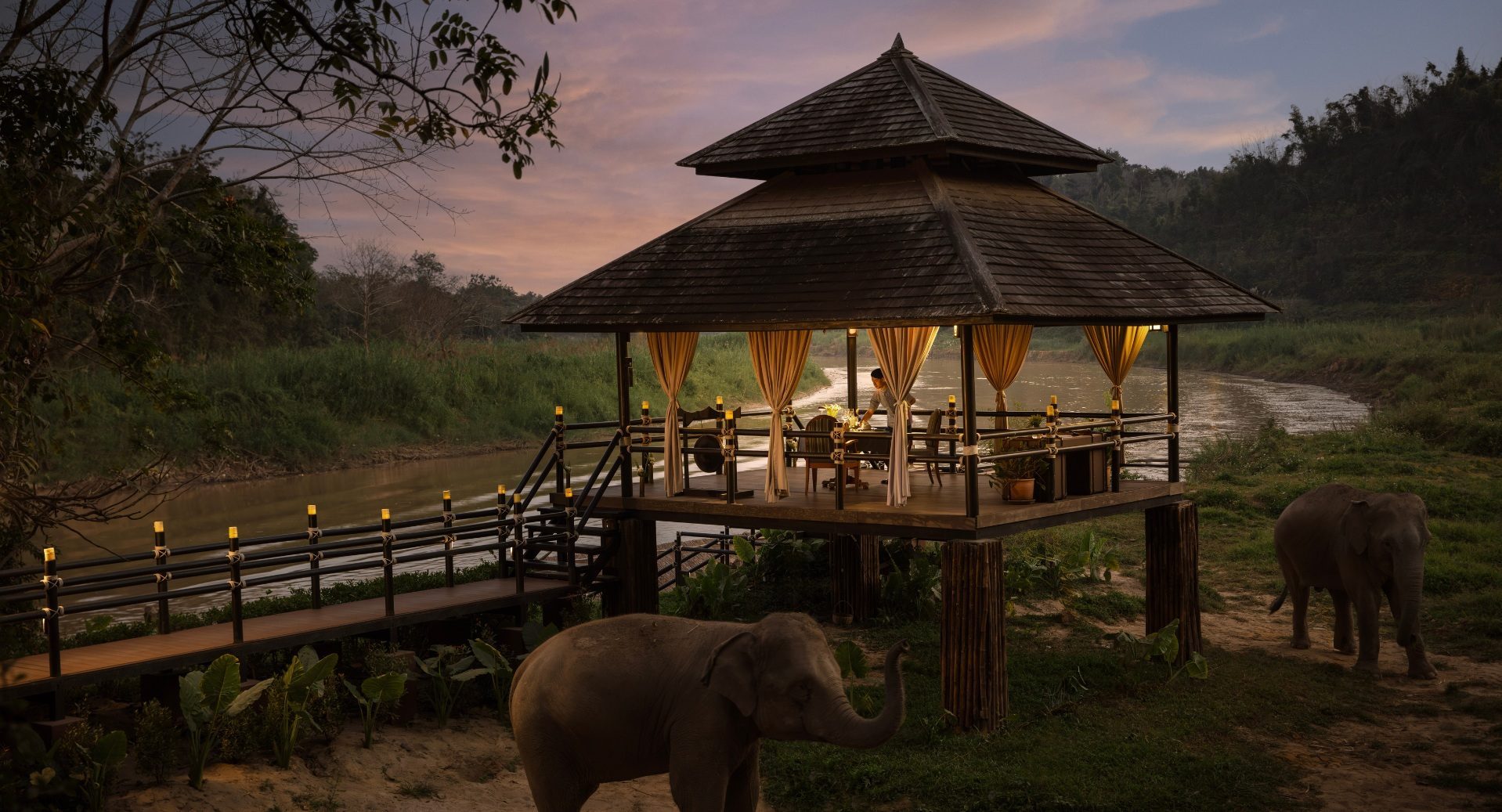 An elevated wooden dining sala on stilts above a river at Anantara Golden Triangle Elephant Camp & Resort at dusk, its curtains lit gold, with two elephants on the riverbank below.