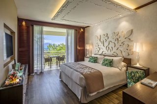 A hotel room with a large bed, a TV, a dining area with a table and chairs, and a balcony with a view of the mountains.
