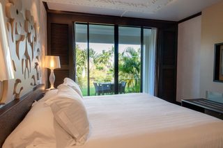 A hotel room with a large bed, a nightstand with a lamp, a view of a garden through a sliding glass door, and a framed picture on the wall.
