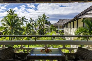 A balcony with wicker furniture and a glass table, overlooking a lush garden with palm trees and a building with a tiled roof.