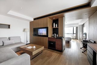 A modern hotel room with a sofa, coffee table, TV, kitchenette, and a view of the city.