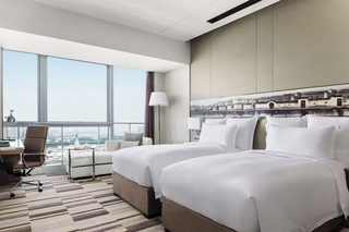A hotel room with two beds, a desk, a chair, a lamp, and a large window offering a city view.