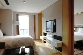 A modern hotel room with a large bed, a flat-screen TV mounted on the wall, a desk with a lamp, and a window with curtains.