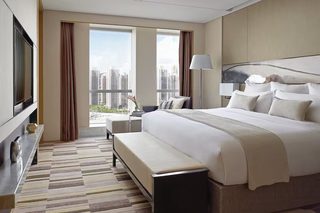 A modern hotel room with a large bed, a flat-screen TV, a bench, and a view of the cityscape.