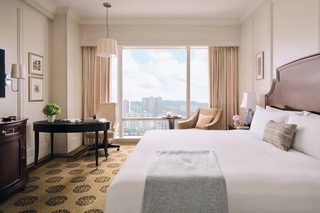 A hotel room with a large bed, a desk with a lamp, a chair, and a view of the cityscape through large windows.