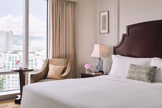 A hotel room with a large bed, a chair, a nightstand, and a window with a city view.