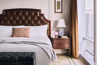 A neatly made bed with a tufted headboard, a bedside table with a lamp and a vase of flowers, and a window with curtains.