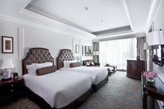 A hotel room with two large beds, a wooden headboard, a desk with a lamp, a dresser, and a large window with curtains.