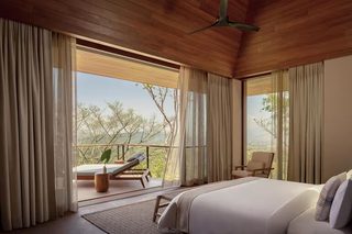 A bedroom with a large bed, a chair, a small table, and a view of trees through a window.