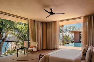 A modern hotel room with a large bed, a ceiling fan, a balcony with a view of the ocean, and a small table with a chair.