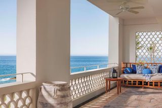 A balcony with a view of the sea, a wooden sofa with cushions, a lantern, and a large vase.