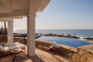 A poolside area with a view of the ocean, a lounge chair, a small table with towels, and a stone wall.