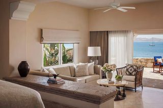 A hotel room with a sofa, armchair, and a dining table. There is a view of the sea through large windows.