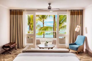 A hotel room with a large window, a ceiling fan, a bed, a coffee table, a blue armchair, and a small table with a vase.
