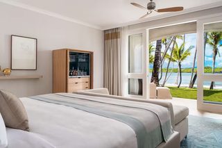 A bedroom with a large bed, a minibar, a ceiling fan, and a view of palm trees and water.