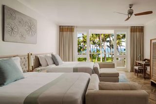 A hotel room with two beds, a desk, and a view of palm trees and the ocean.