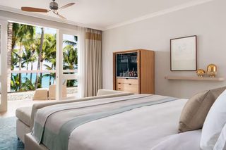 A hotel room with a large bed, a seating area with a view of palm trees, a minibar, and a ceiling fan.