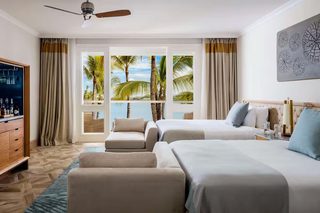 A hotel room with two beds, a sofa, a TV, and a view of palm trees and the sea.
