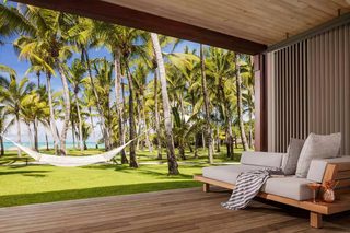 A wooden deck with a hammock, a sofa with cushions, and a view of palm trees and the ocean.