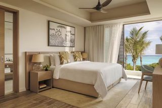 A bedroom with a large bed, a nightstand with a lamp, a framed picture on the wall, and a view of the ocean through the window.