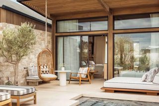 A modern outdoor pool area with a stone wall, wooden deck, and seating including a wicker chair and a cushioned bench.