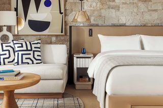 A bedroom with a bed, a nightstand, a lamp, a chair, a round table, and a stone wall.