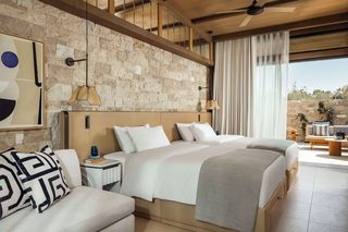 A modern hotel room with a stone wall, a large bed, a sofa, and a dining area with a view.