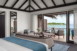 A bedroom with a large bed, a sitting area with a table and chairs, and a balcony with a view of the water.