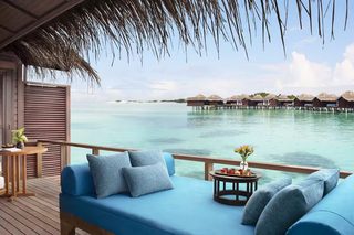 A wooden deck with a blue sofa and cushions, a small table with a vase of flowers, and a view of overwater bungalows and calm sea.