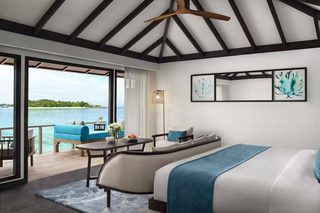 A modern bedroom with a large bed, a sofa, and a dining table. There is a view of the sea and a deck outside.