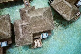 An aerial view of overwater bungalows with thatched roofs, clear turquoise water, and wooden walkways.