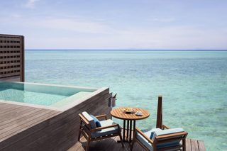 A wooden deck with a glass infinity pool, lounge chairs, and a table with a drink and snacks.