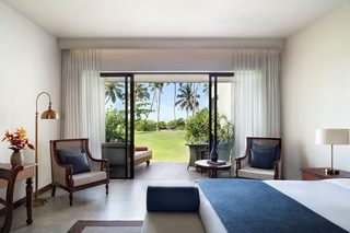 A hotel room with a large bed, two armchairs, a small table, and a view of a tropical garden through a sliding glass door.