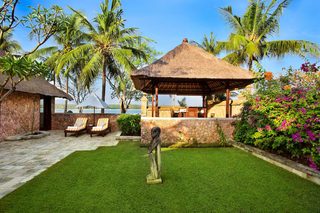 A thatched-roof gazebo, a stone wall, and a statue in a well-maintained garden with palm trees and flowering plants.