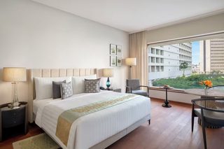 A hotel room with a large bed, two bedside tables with lamps, a chair, a small table with flowers, and a view of buildings and greenery outside the window.