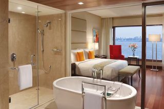 A modern hotel room with a large bathtub, a separate shower area, a bed with a view of the city, and a red chair.