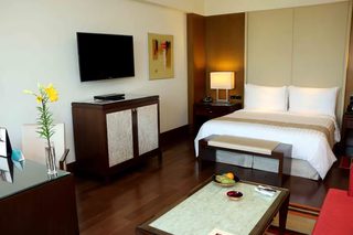 A hotel room with a bed, bedside table, TV, artwork, and a desk with a vase of flowers.