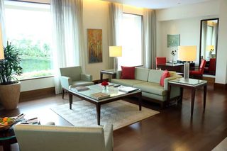 A modern living room with a sofa, armchairs, a coffee table, and a dining table. There is a large window with curtains, a potted plant, and artwork on the walls.