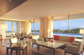 A hotel room with a large window, a sofa, a coffee table, and a dining area with chairs and a table.