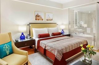 A hotel room with a bed, bedside tables, a chair, a glass-enclosed bathroom, and artwork on the wall.