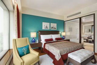 A hotel room with a large bed, two bedside tables, a chair, a window with curtains, and a bathroom visible through a glass door.