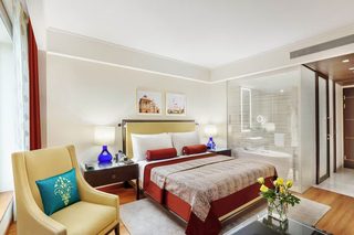 A hotel room with a bed, a chair, a nightstand, a lamp, and a bathroom with a glass shower enclosure.