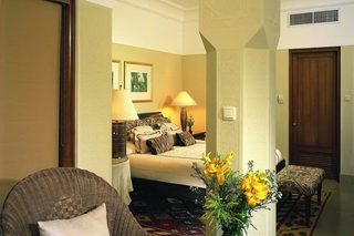 A hotel room with a bed, a sitting area with a chair and a table, a vase with flowers, and a door.