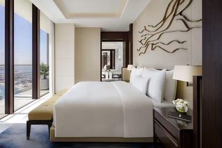 A modern hotel room with a large bed, a bedside table with a lamp, a view of the city, and a decorative wall piece.